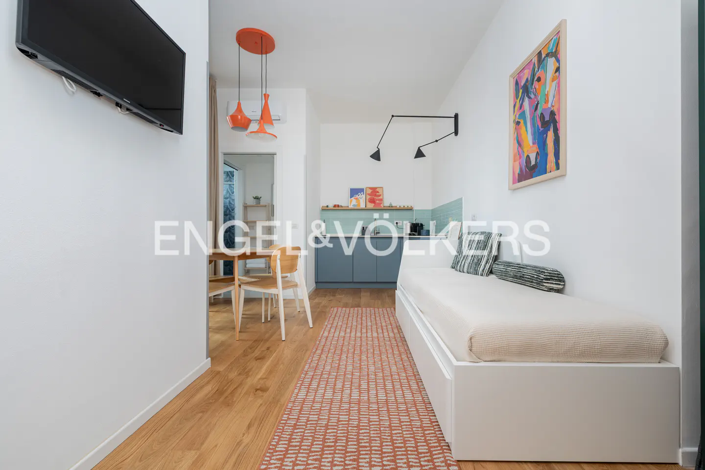 Modern studio apartment with a daybed, patterned rug, dining area, and kitchenette with blue cabinets and orange lighting.
