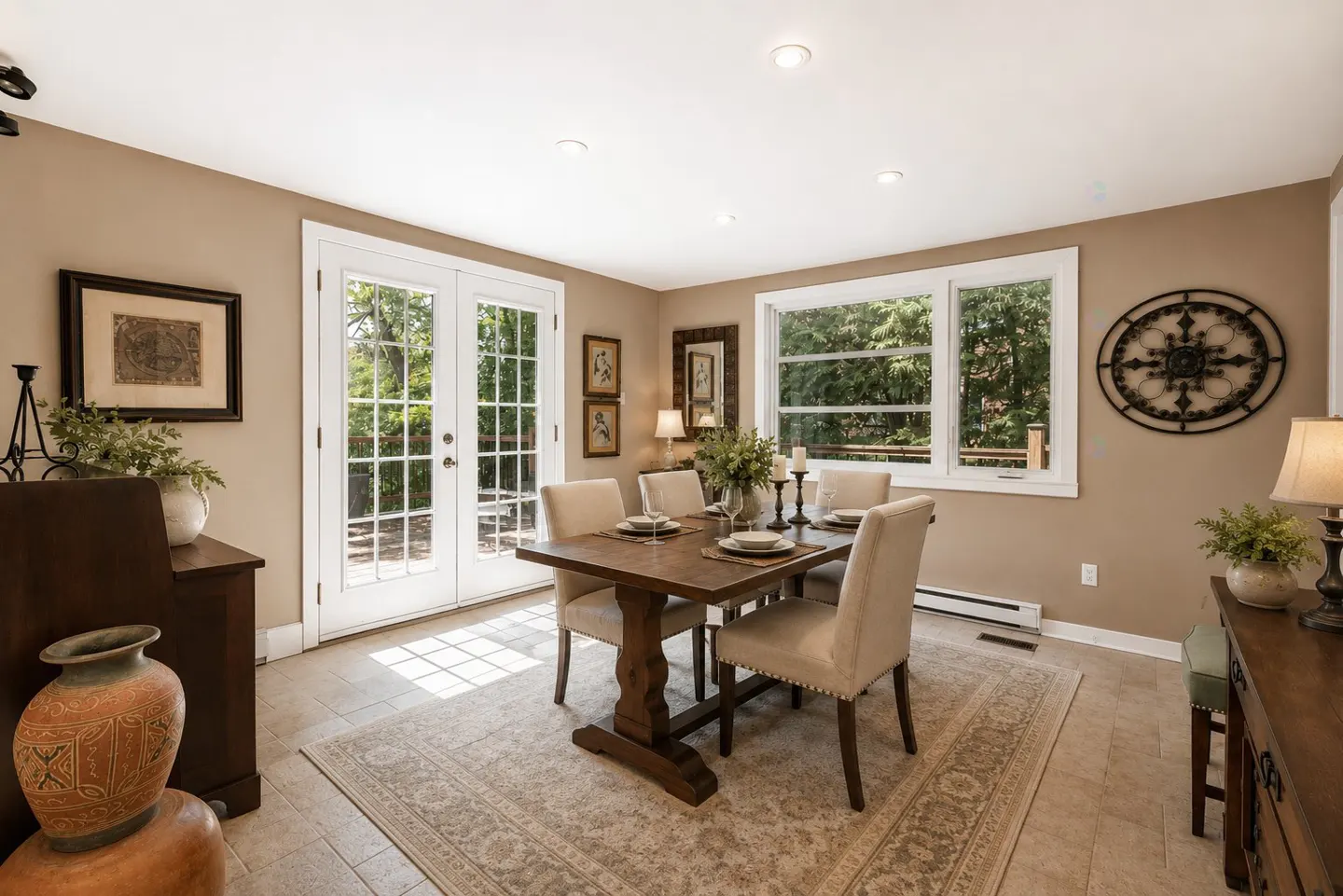 Dining room with a wooden table, four beige chairs, a patterned rug, French doors, and windows overlooking greenery.