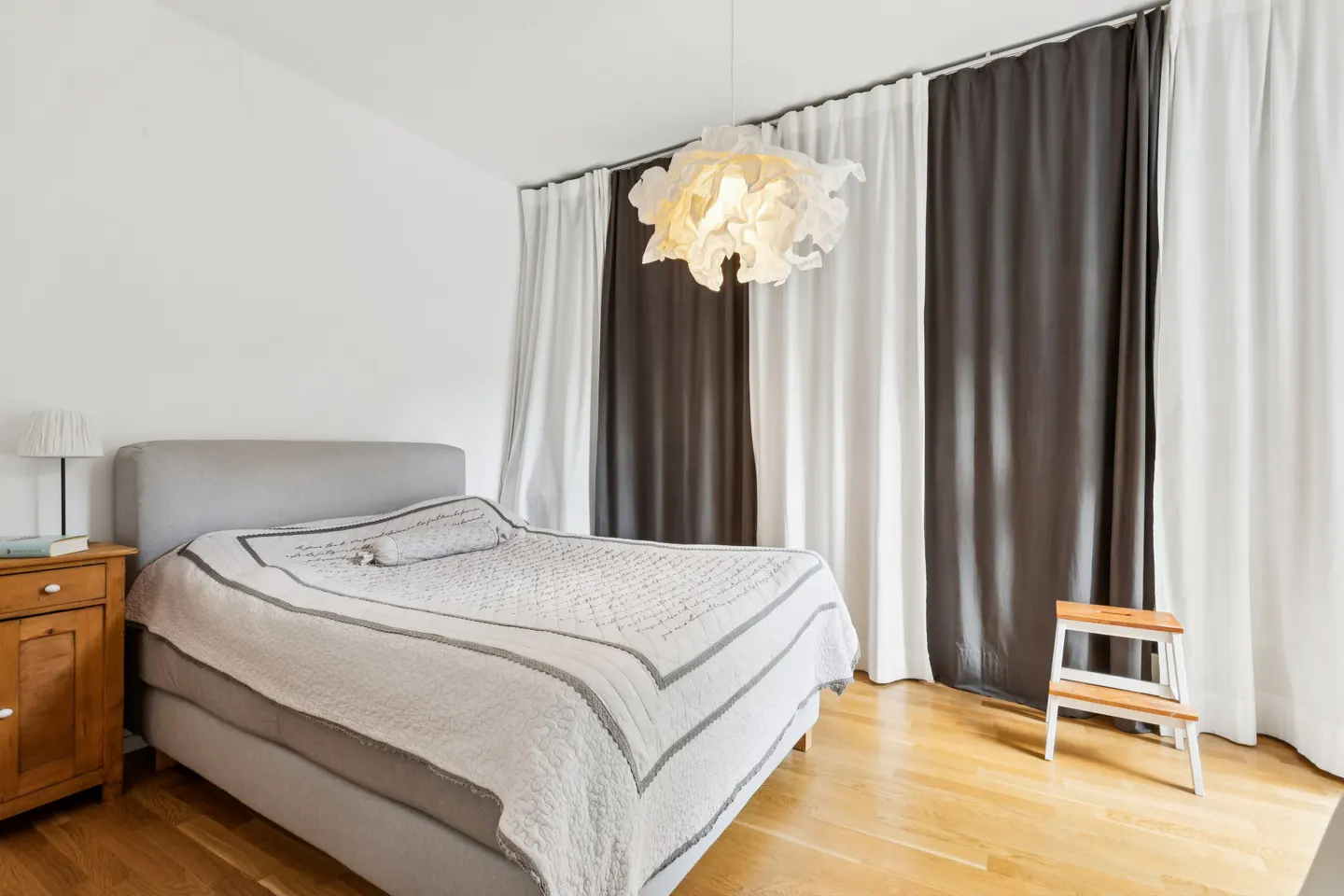 Bedroom with a gray bed, white and gray quilt, wood floors, and white and gray curtains. A modern light fixture hangs from the ceiling.