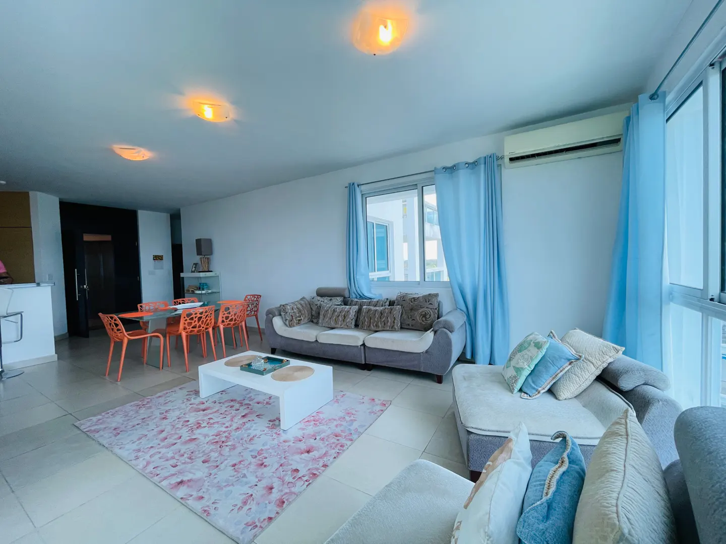 Bright living room with sofas, a white coffee table on a floral rug, and an orange dining set near a dark door. Blue curtains frame the window.