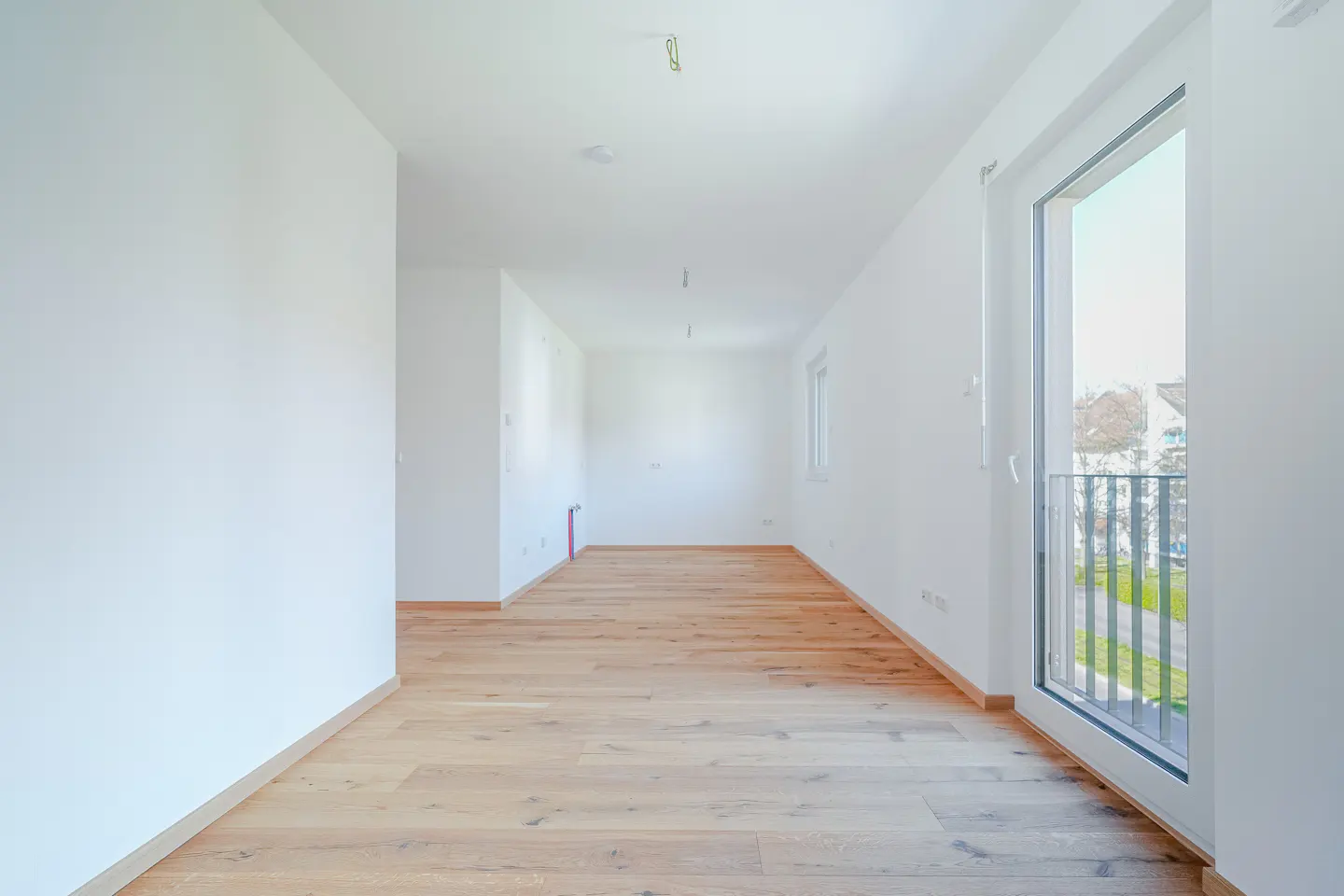 Bright, empty room with light wood floors and white walls. A sliding glass door leads to a balcony with a view of greenery.