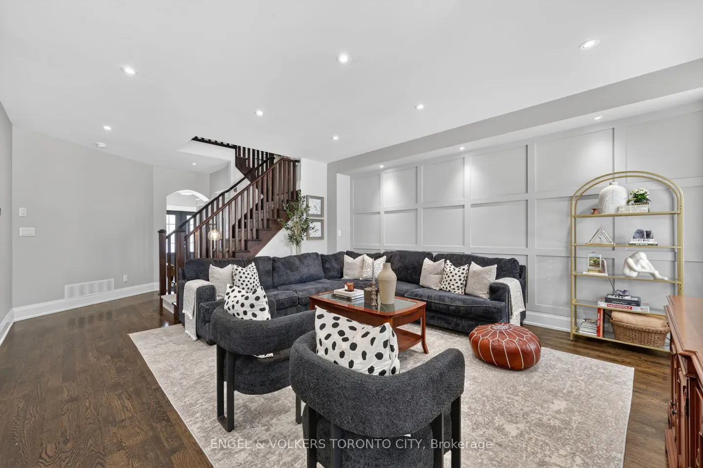 Living room with dark wood floors, gray walls, and a large gray sectional sofa with decorative pillows. Two gray chairs face the sofa. A staircase is visible in the background.