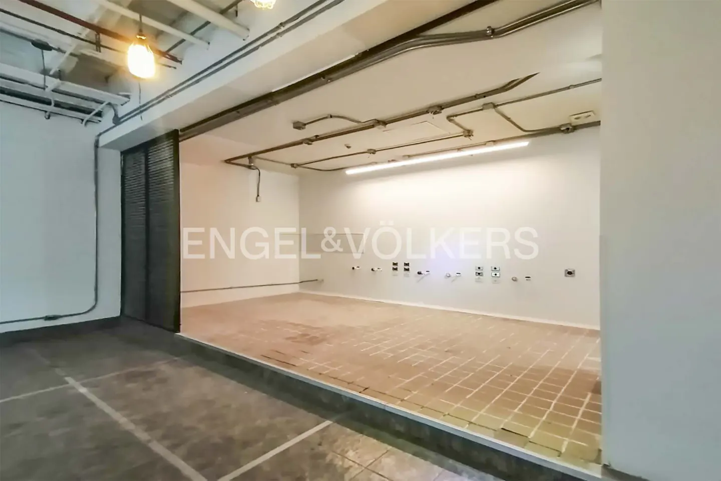 Empty white room with brick floor and Engel & Völkers logo on the wall. Metal pipes run along the ceiling.