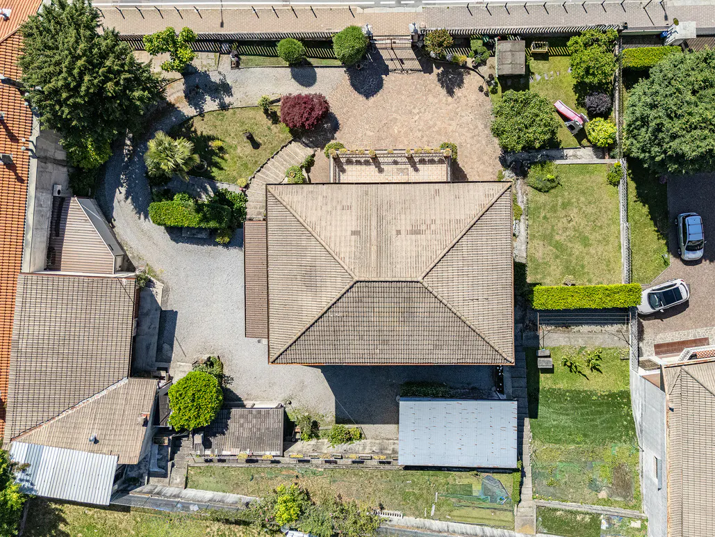 Aerial view of a house with a tiled roof, surrounding gravel driveway, green lawns, trees, and two parked cars.