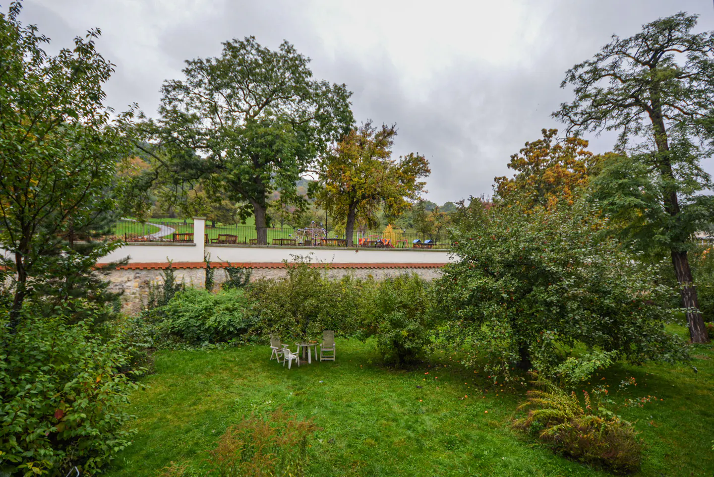 A green lawn with a white table and chairs, trees, and a white wall with a park in the background.
