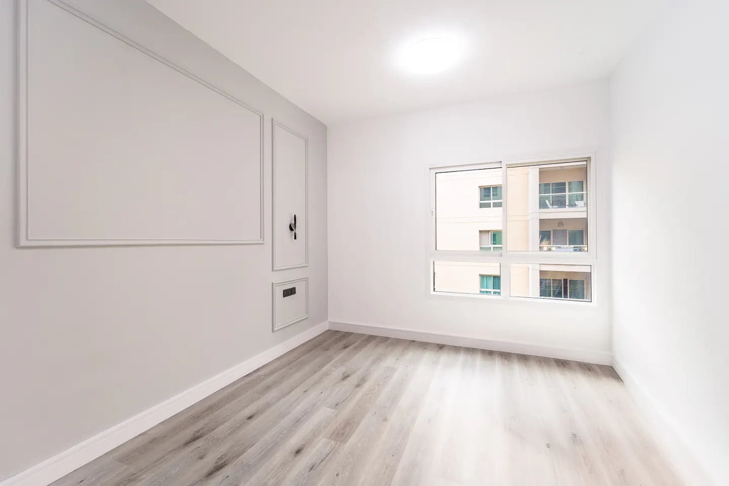 Bright, empty room with gray wood floors, white walls, and a window showing a building exterior.