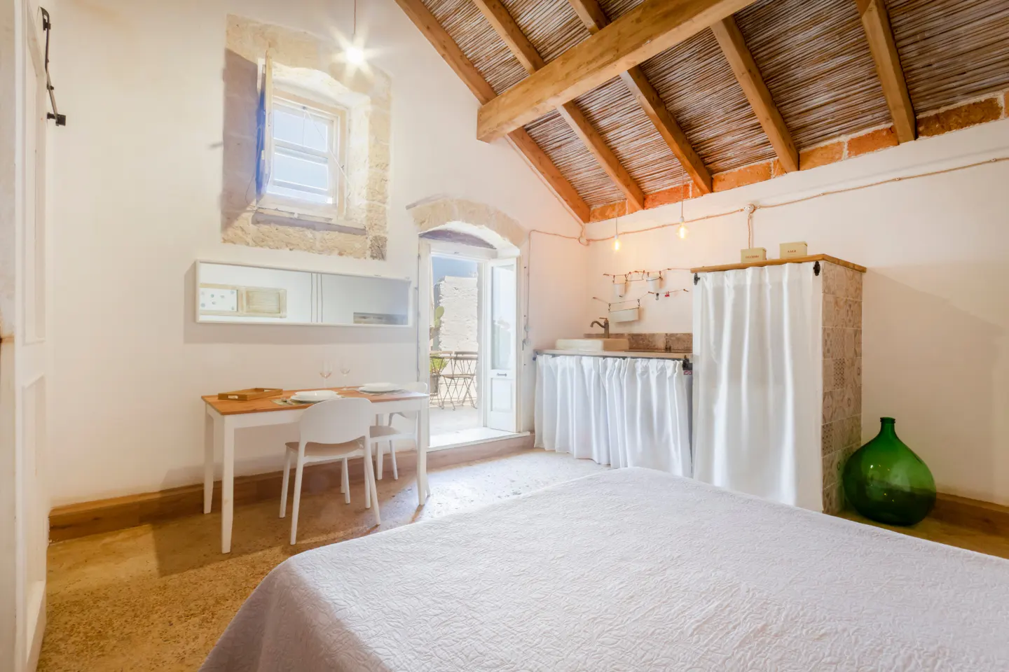 Rustic bedroom with a bed, small dining table, kitchenette with white curtains, and a vaulted wooden ceiling.