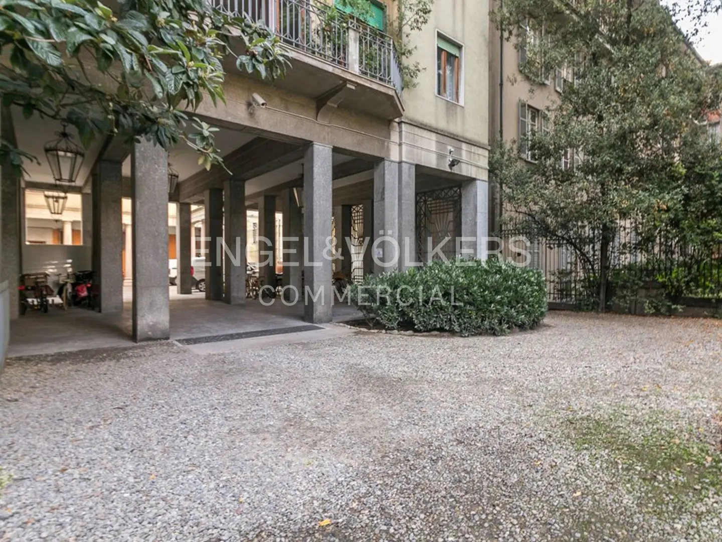 Exterior view of a building with a covered walkway supported by gray pillars, a gravel courtyard, and green foliage.