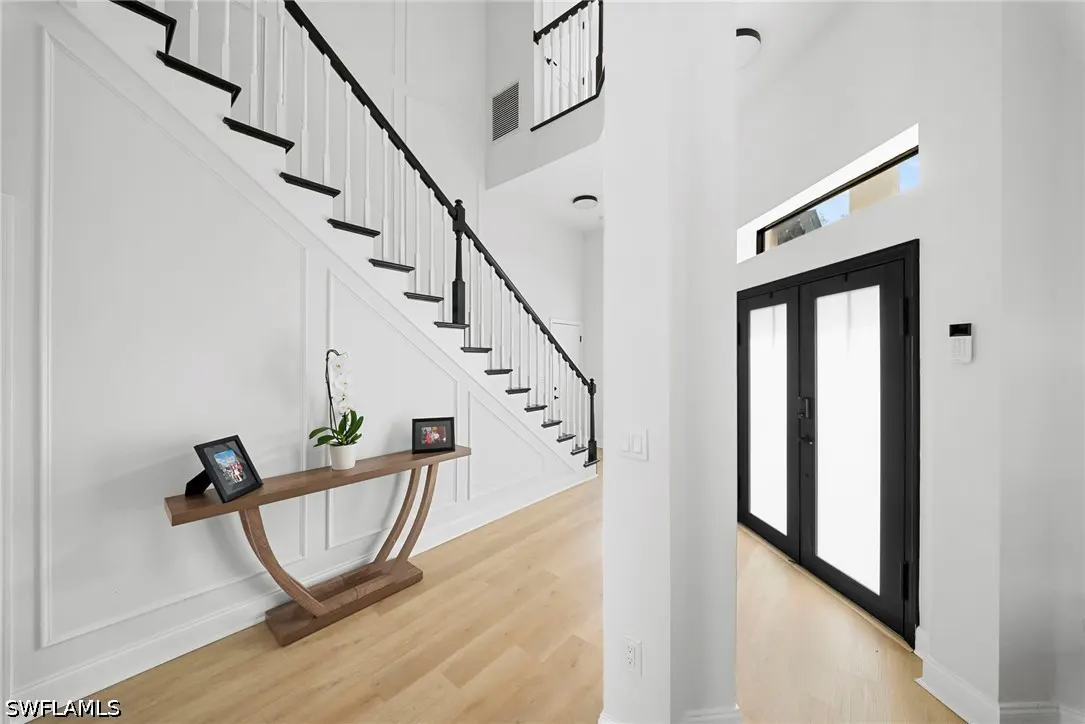 Bright foyer with white walls, light wood floors, and black accents. A staircase and console table are on the left, and a black double door is on the right.