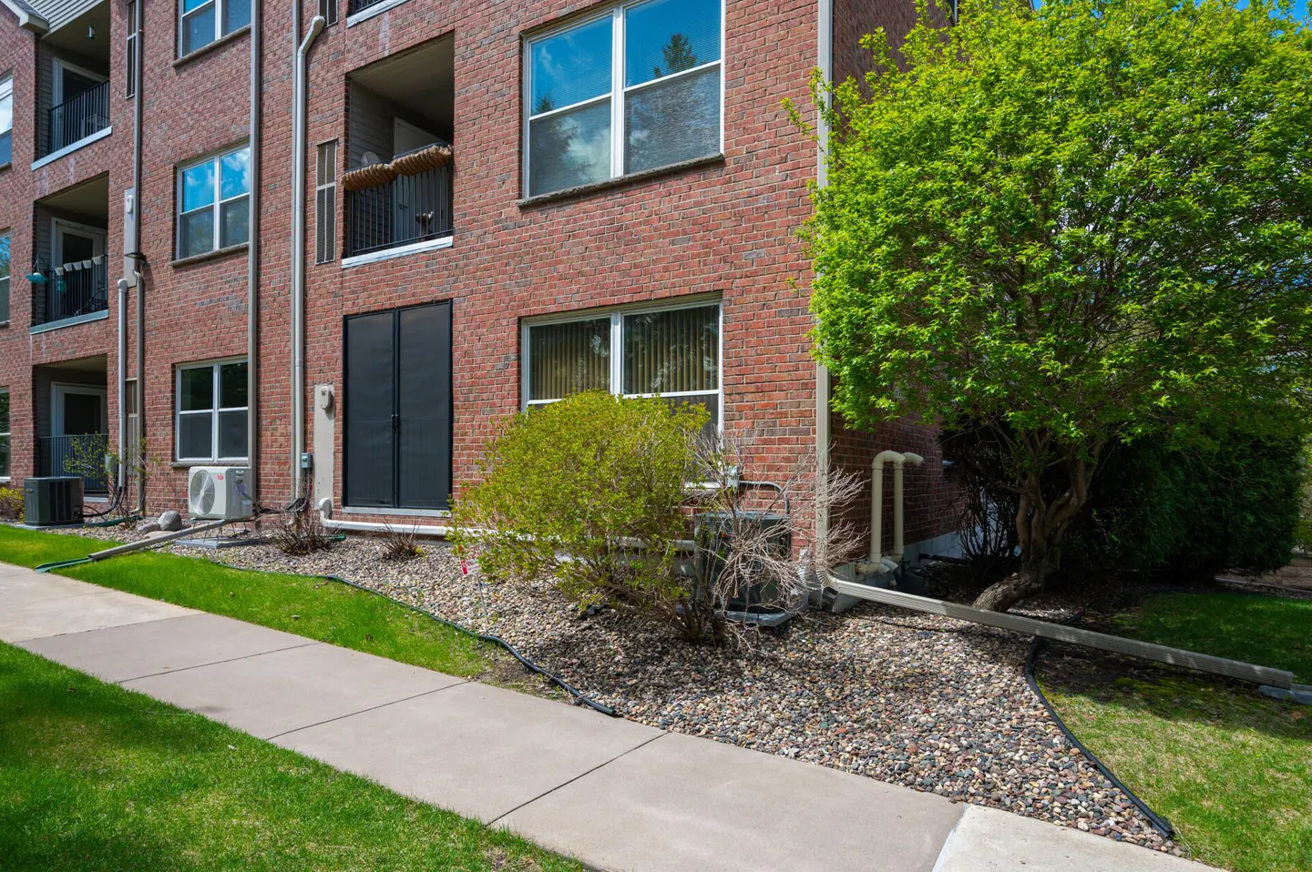 Brick apartment building with black doors, windows, and balconies. Green grass and trees surround the building. A concrete sidewalk runs along the front.