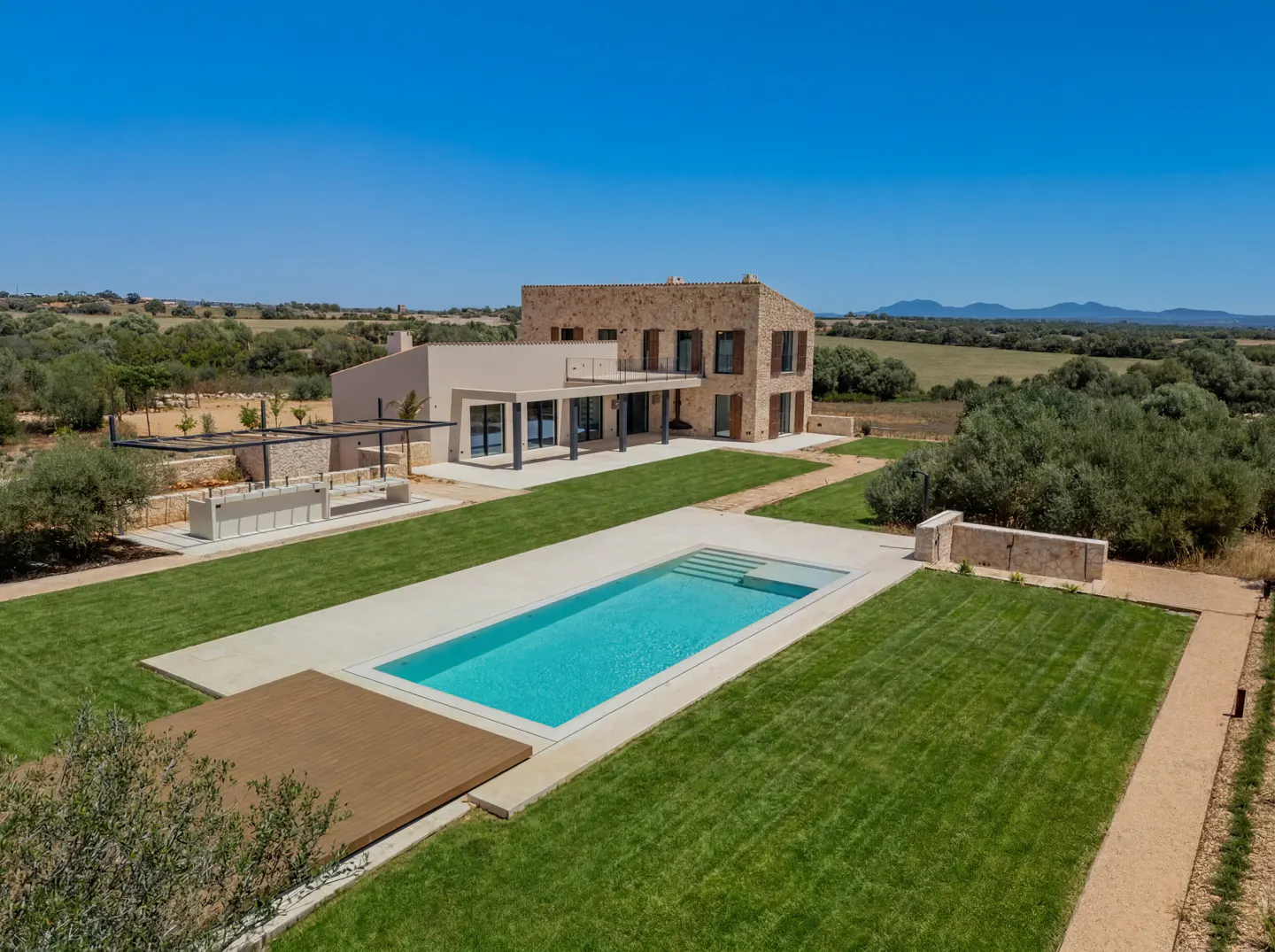 Modern stone villa with a swimming pool, wooden deck, and lush green lawn set in a rural landscape under a clear blue sky.