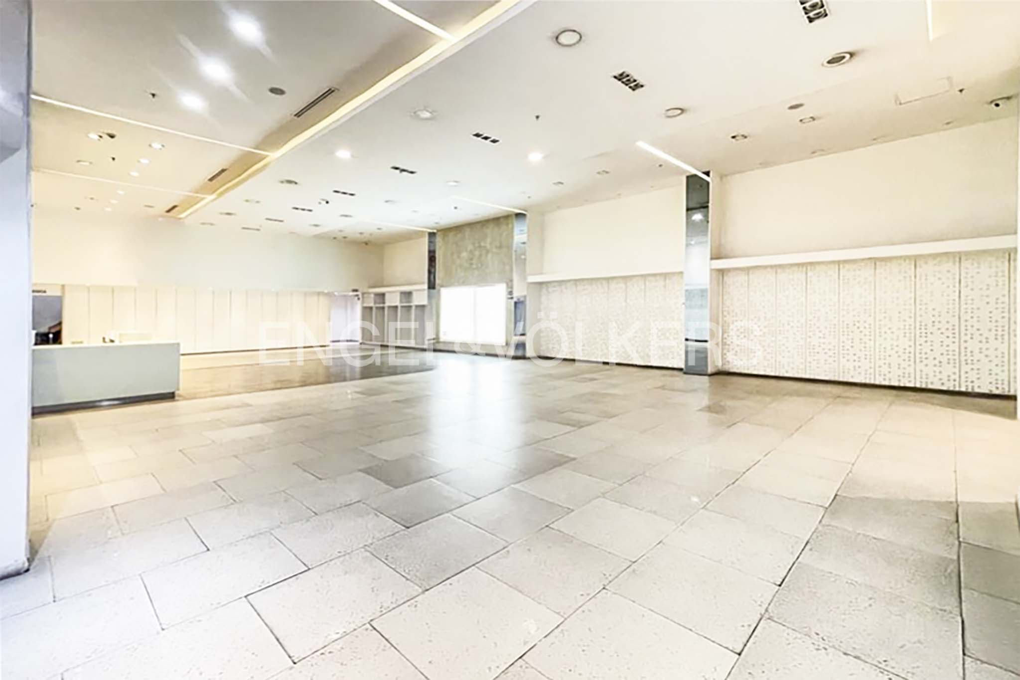 Bright, empty commercial space with white walls, tile floors, and recessed lighting. A reception desk and shelving are visible.