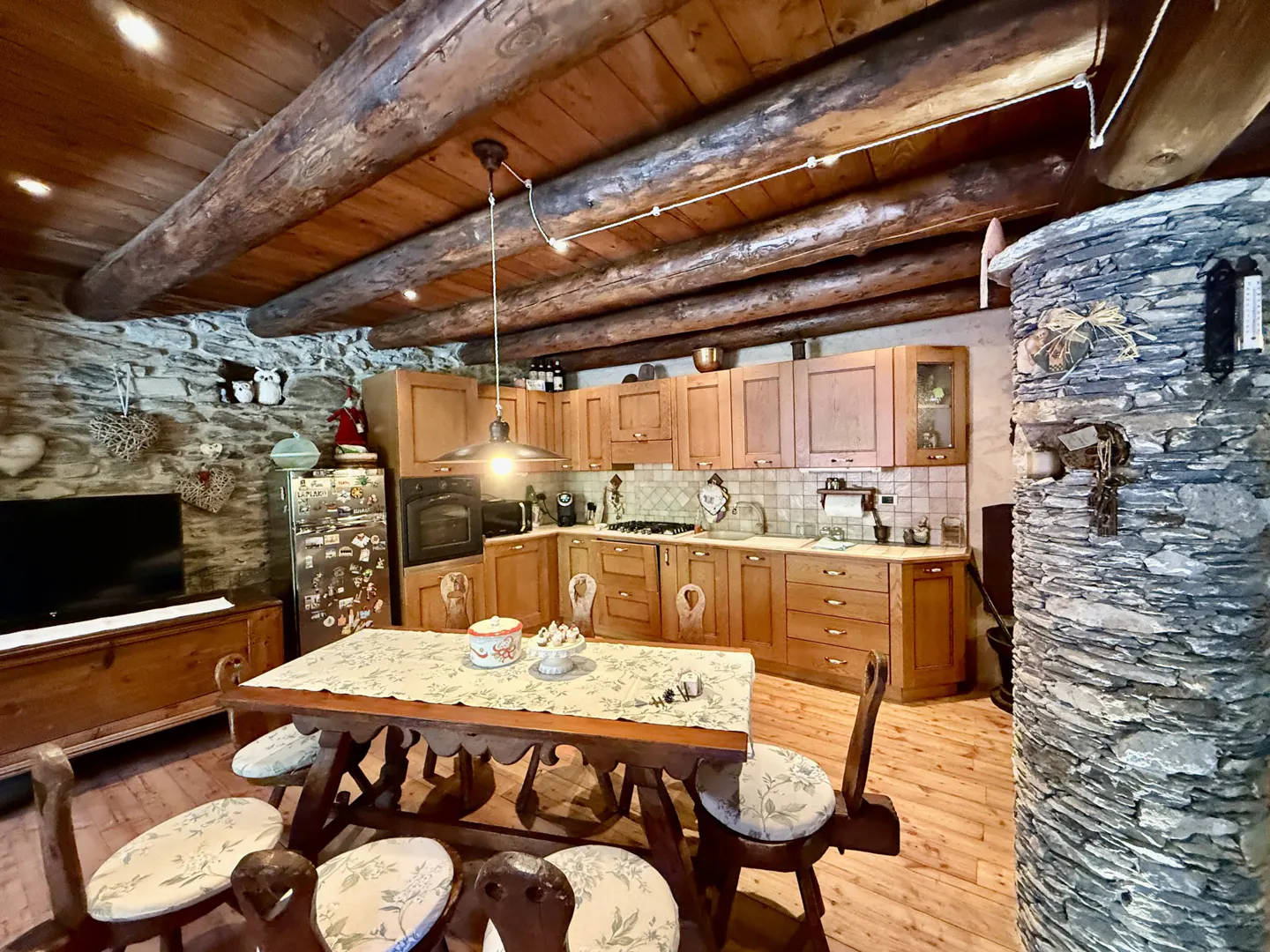 A rustic kitchen with wood cabinets, a stone wall, and a wooden table with chairs. The ceiling has exposed wooden beams.