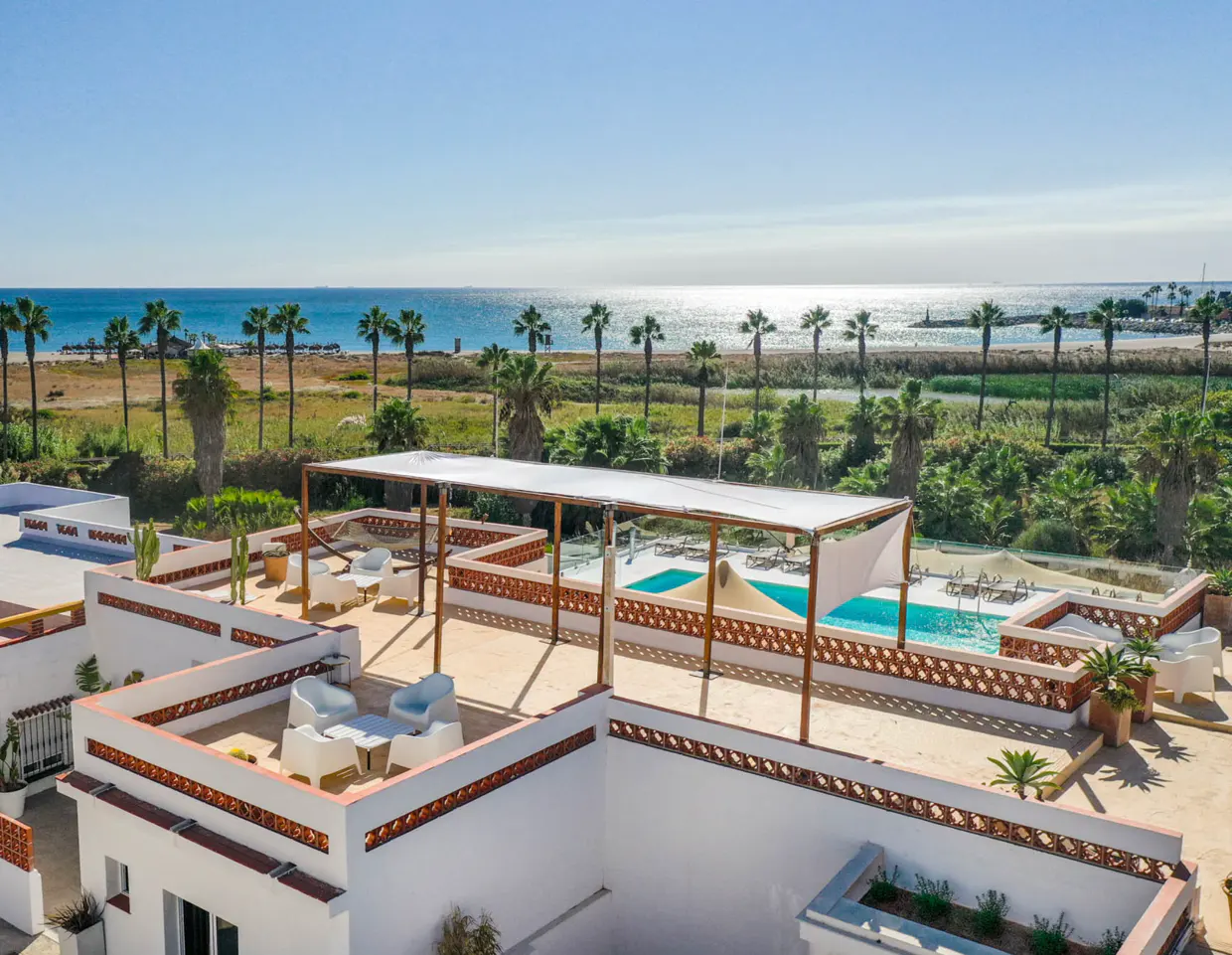 Rooftop view of a white building with a pool and lounge area, overlooking the ocean and palm trees on a sunny day.