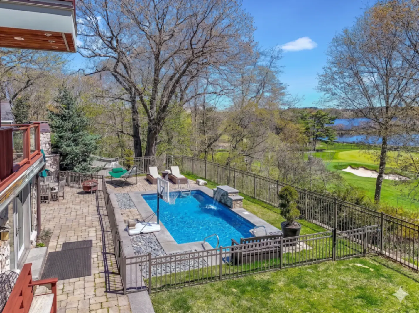 Aerial view of a backyard with a blue pool, lounge chairs, and a patio, fenced in with a golf course in the background.