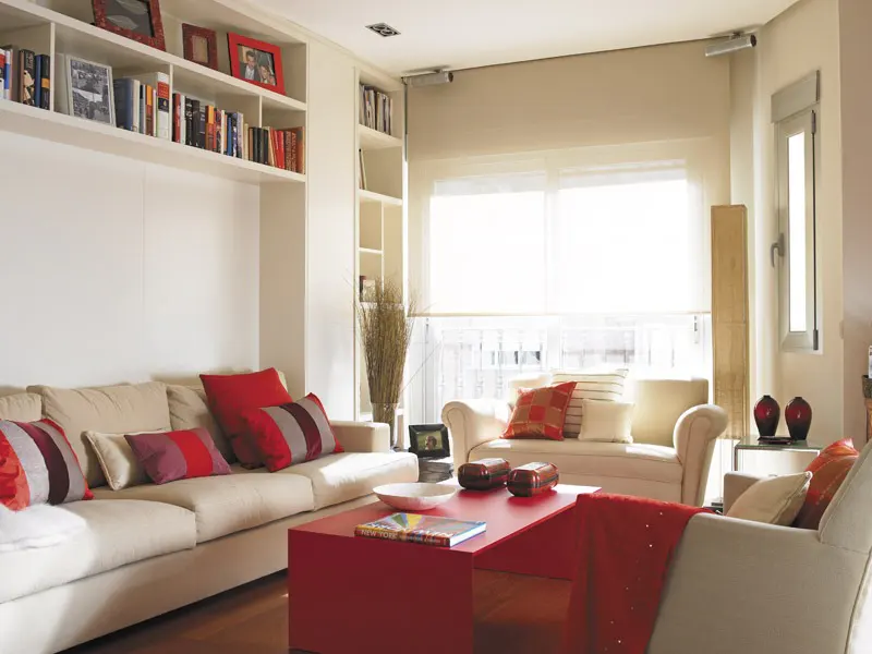 Bright living room with white walls, built-in bookshelves, and light beige sofas with red accents. A red coffee table sits in the center.
