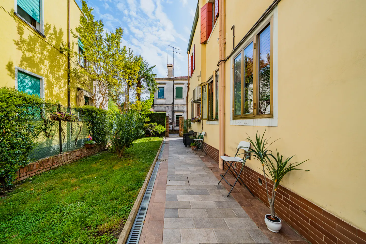 A narrow walkway between yellow buildings leads to a distant building under a blue sky. A green lawn and plants line the walkway.