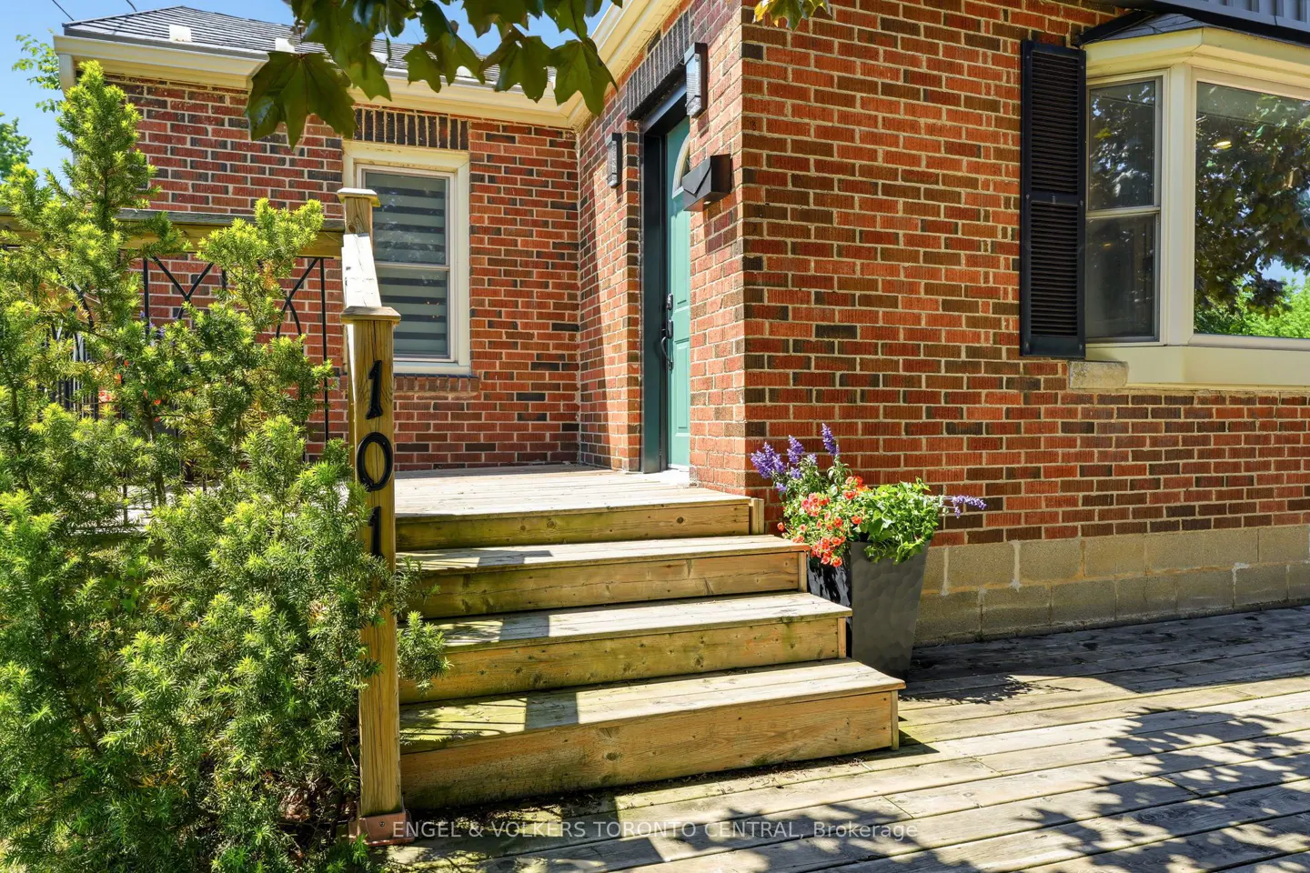 Red brick house exterior with wooden steps leading to a teal front door, featuring a potted plant and lush greenery.