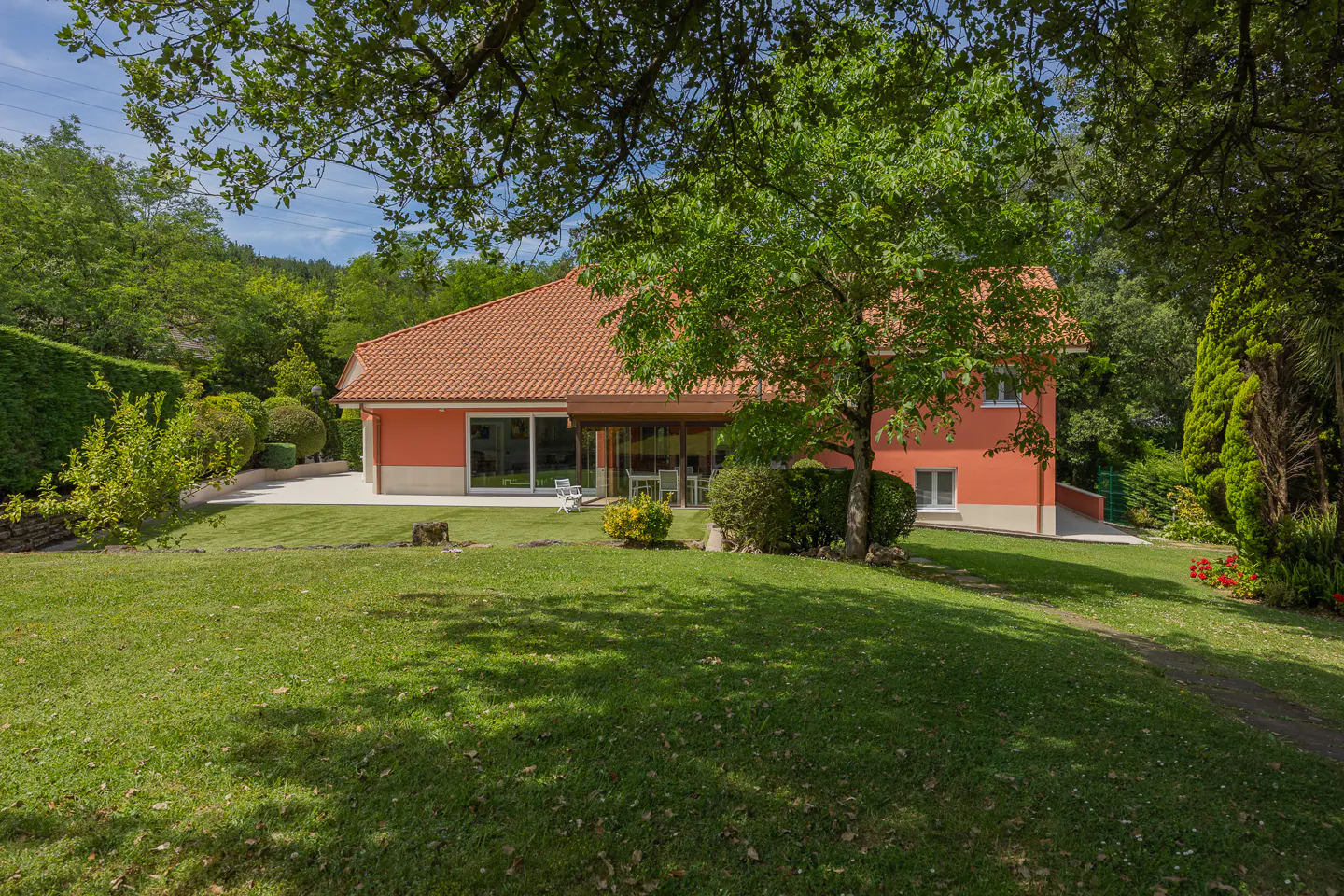 A peach-colored house with a terracotta roof sits in a lush green garden surrounded by trees on a sunny day.