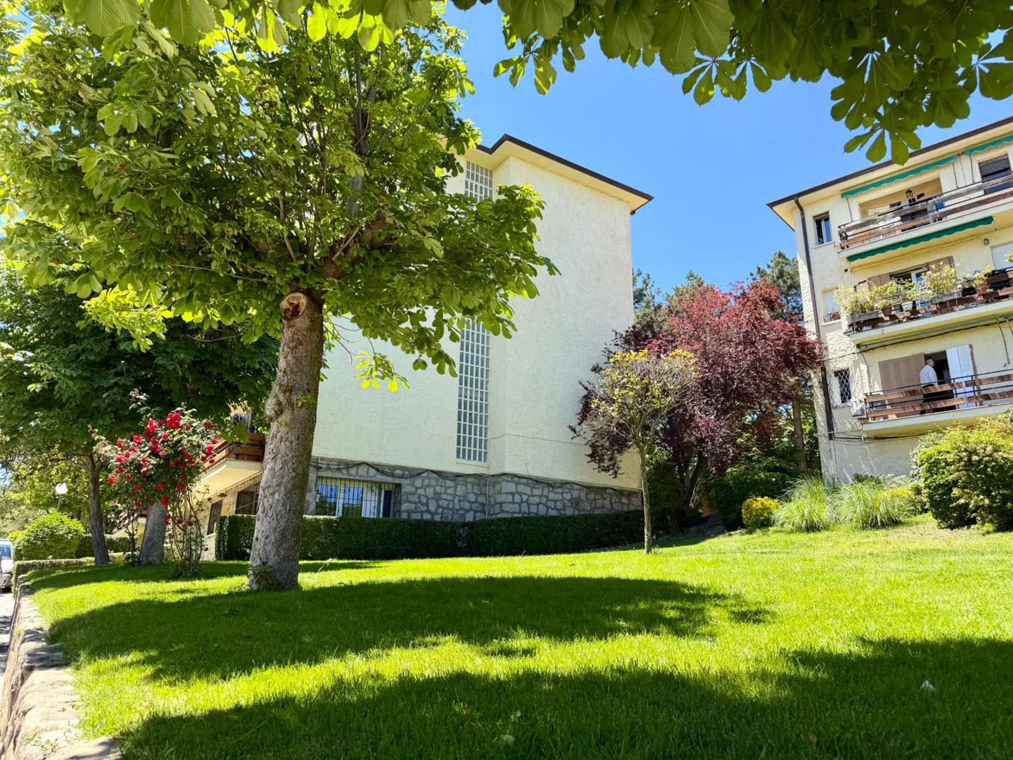 Exterior view of a light yellow apartment building with balconies, surrounded by green trees and lawn on a sunny day.