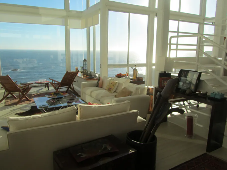 Bright living room with ocean view. White sofas, chairs, and large windows overlooking the sea. A staircase is visible on the right.