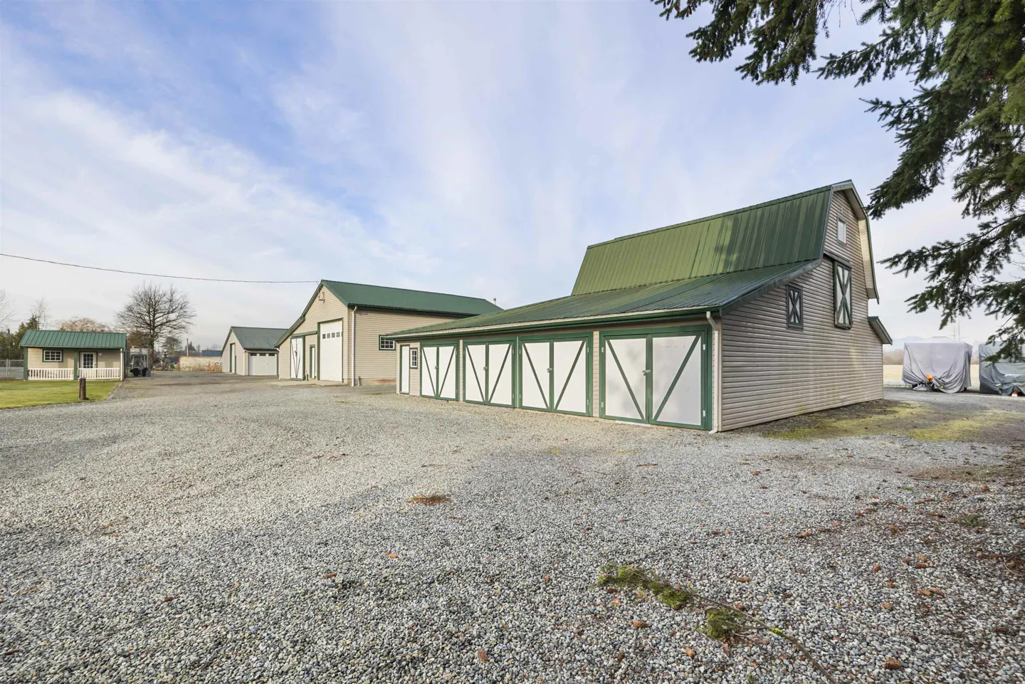A gravel lot leads to a barn with green roof and white doors, and other outbuildings.