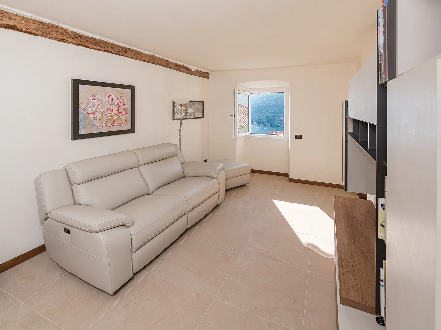 Living room with a beige leather sofa, ottoman, and a view of a lake through an open window. A modern bookshelf is on the right.