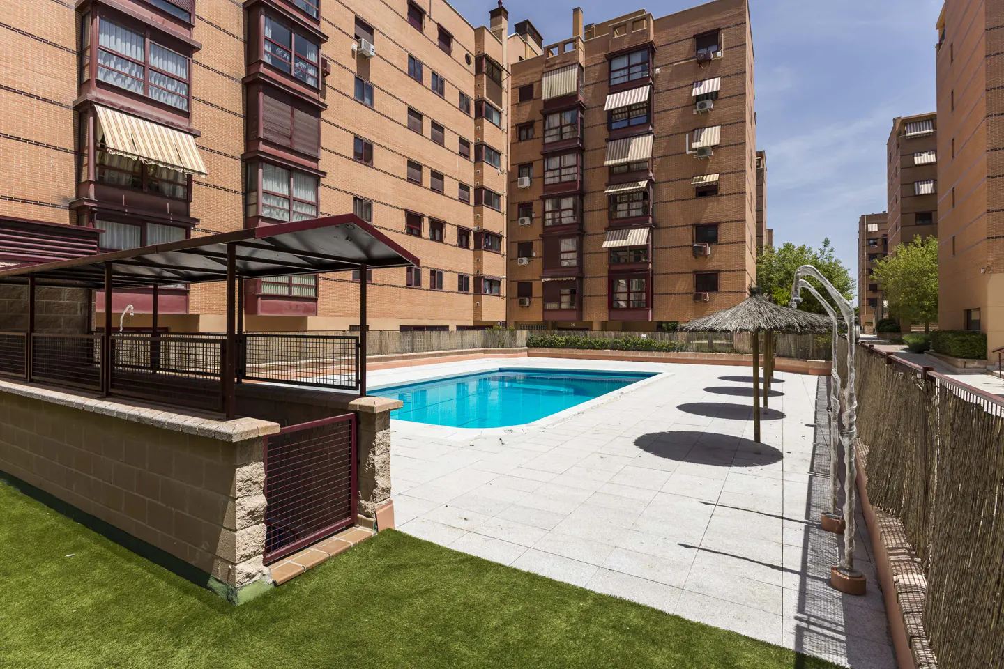 Apartment complex with a pool. Brick buildings surround a blue swimming pool with a covered seating area and straw umbrellas.