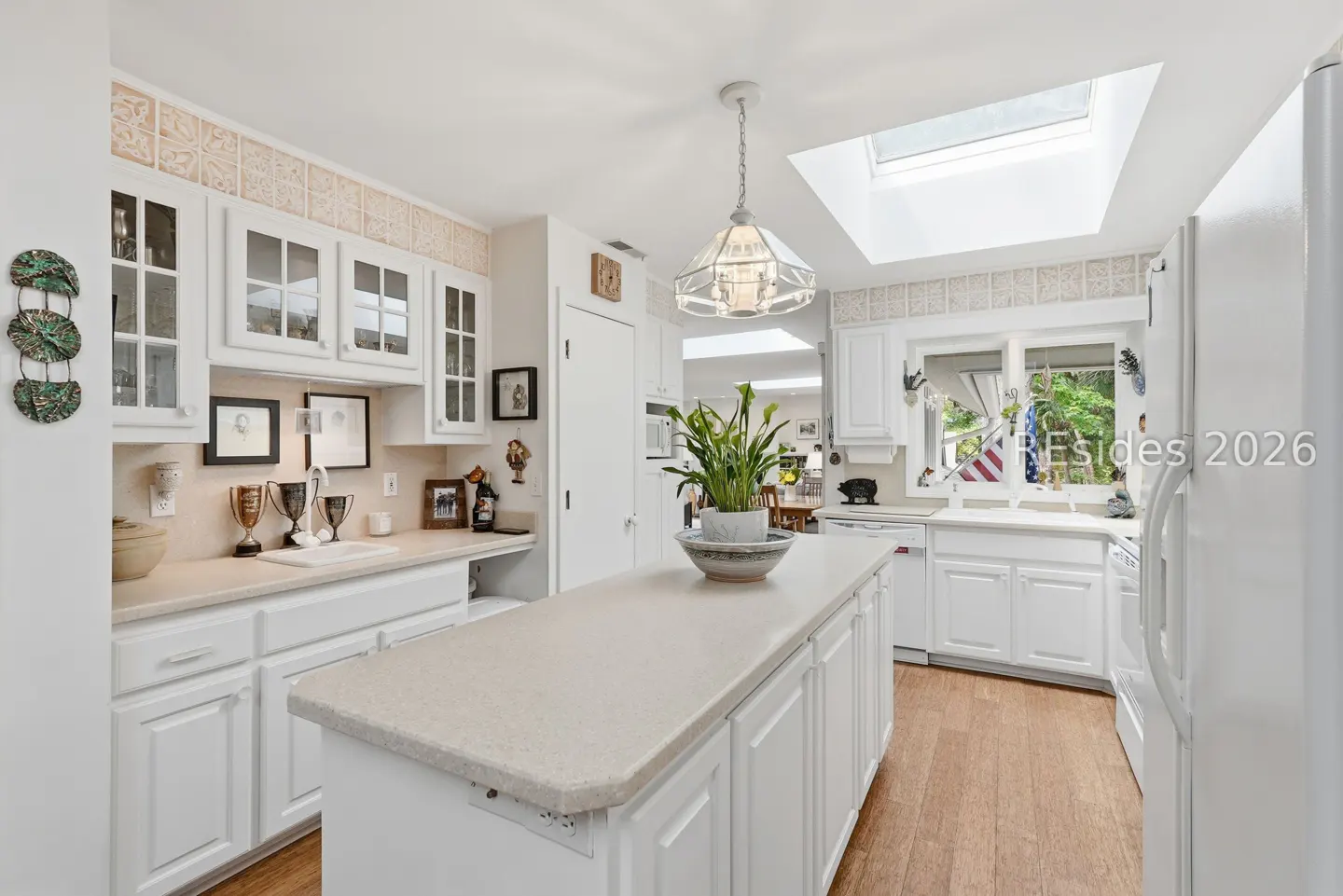 Bright kitchen with white cabinets, countertops, and island. A skylight provides natural light. Wood floors add warmth. A plant sits on the island.