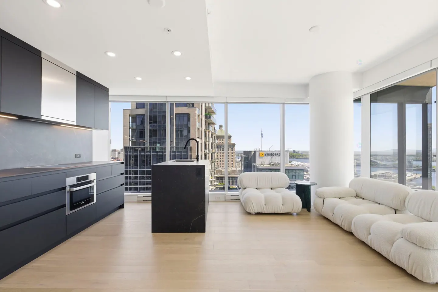 Bright, modern condo interior with black kitchen cabinets, island, and white modular sofa. Large windows offer city views.