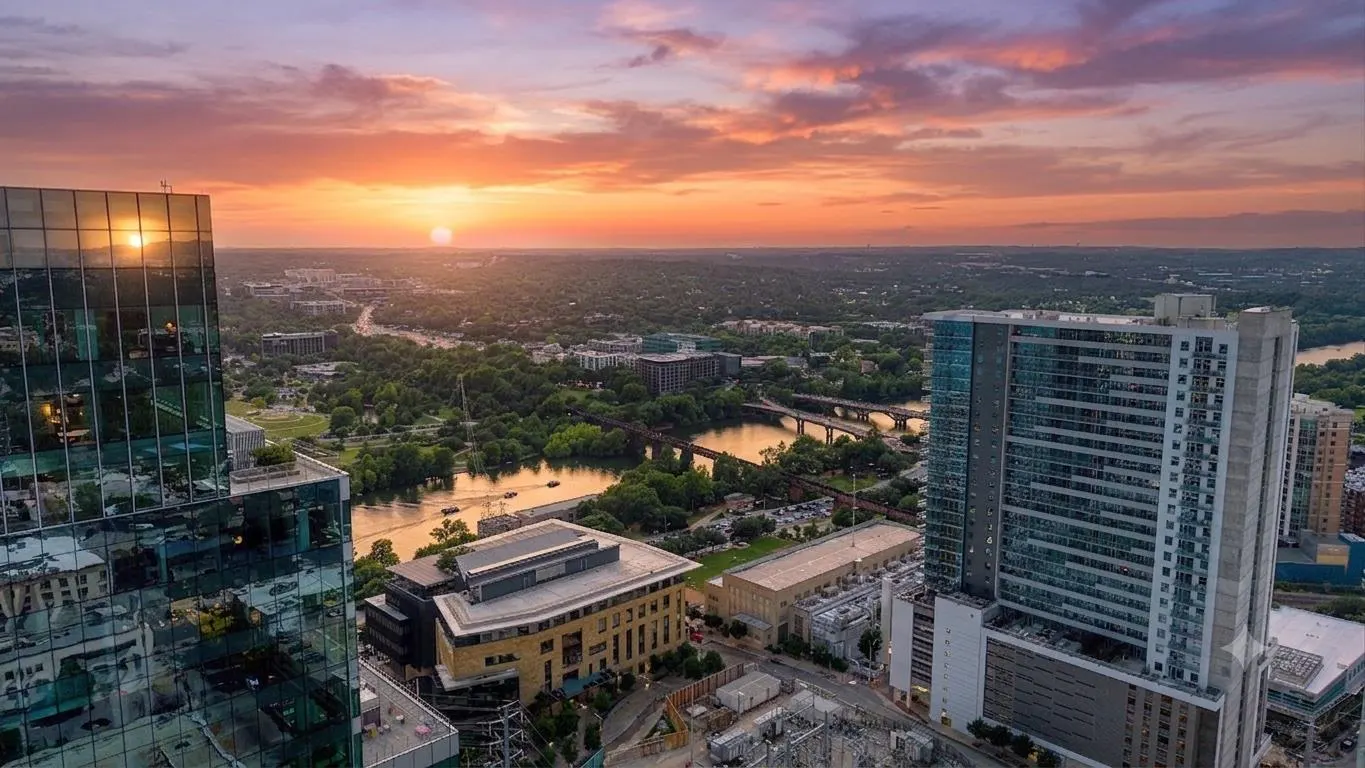 Aerial view of a city skyline at sunset, featuring modern glass skyscrapers, a river, and lush greenery under a pink sky.
