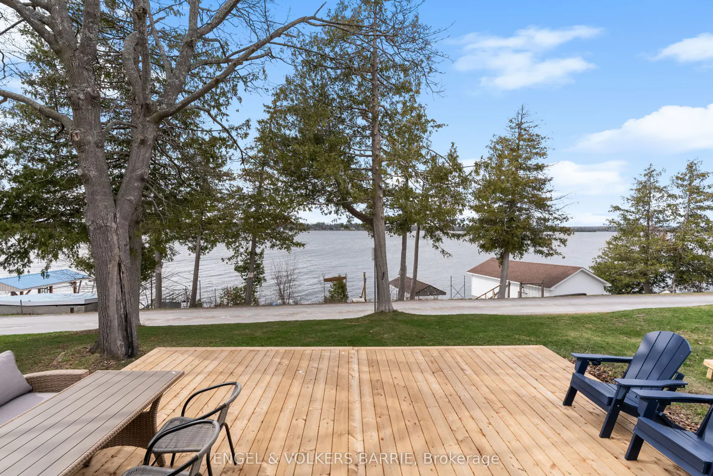 Wooden deck with blue Adirondack chairs and a table overlooks a lake with trees and houses in the background under a blue sky.