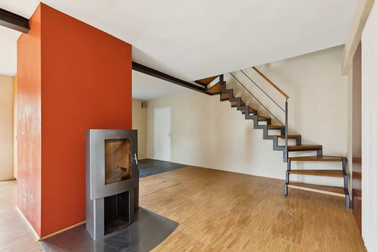 Bright living room with wood floors, orange accent wall, and modern metal fireplace. Open staircase with metal railings leads to the upper level.
