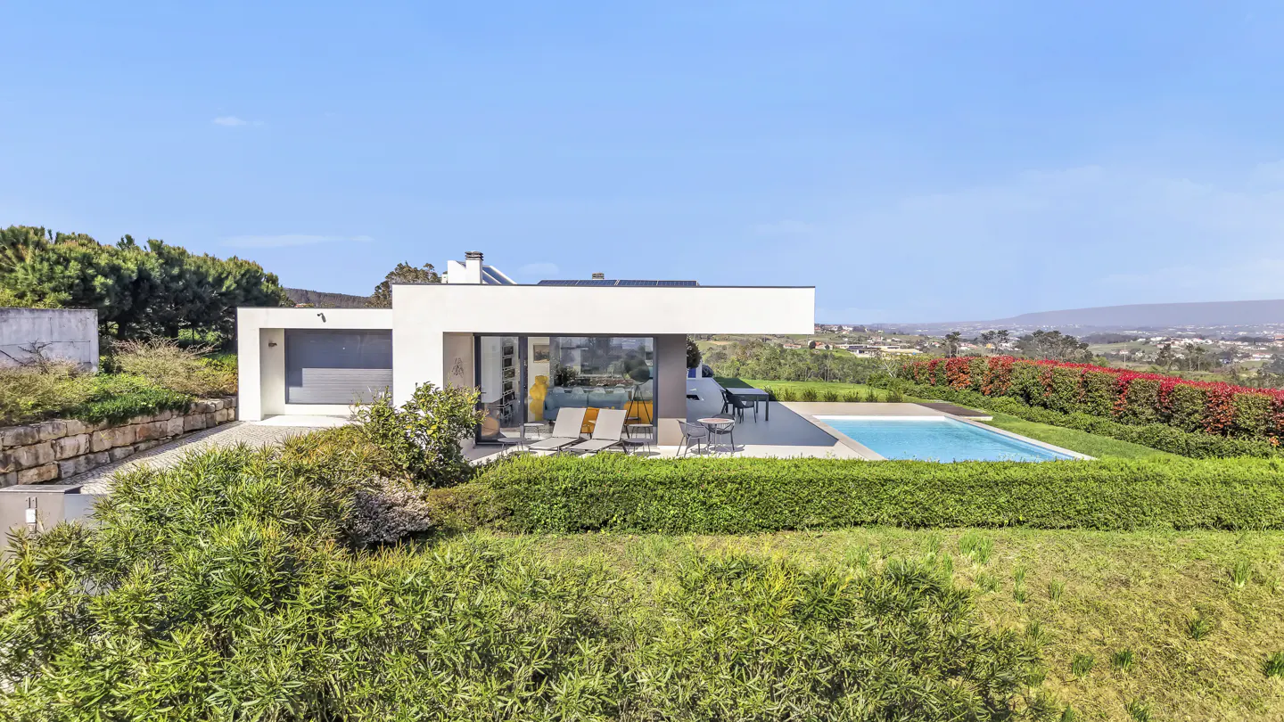 Modern white house with a pool, surrounded by green bushes and trees under a blue sky.