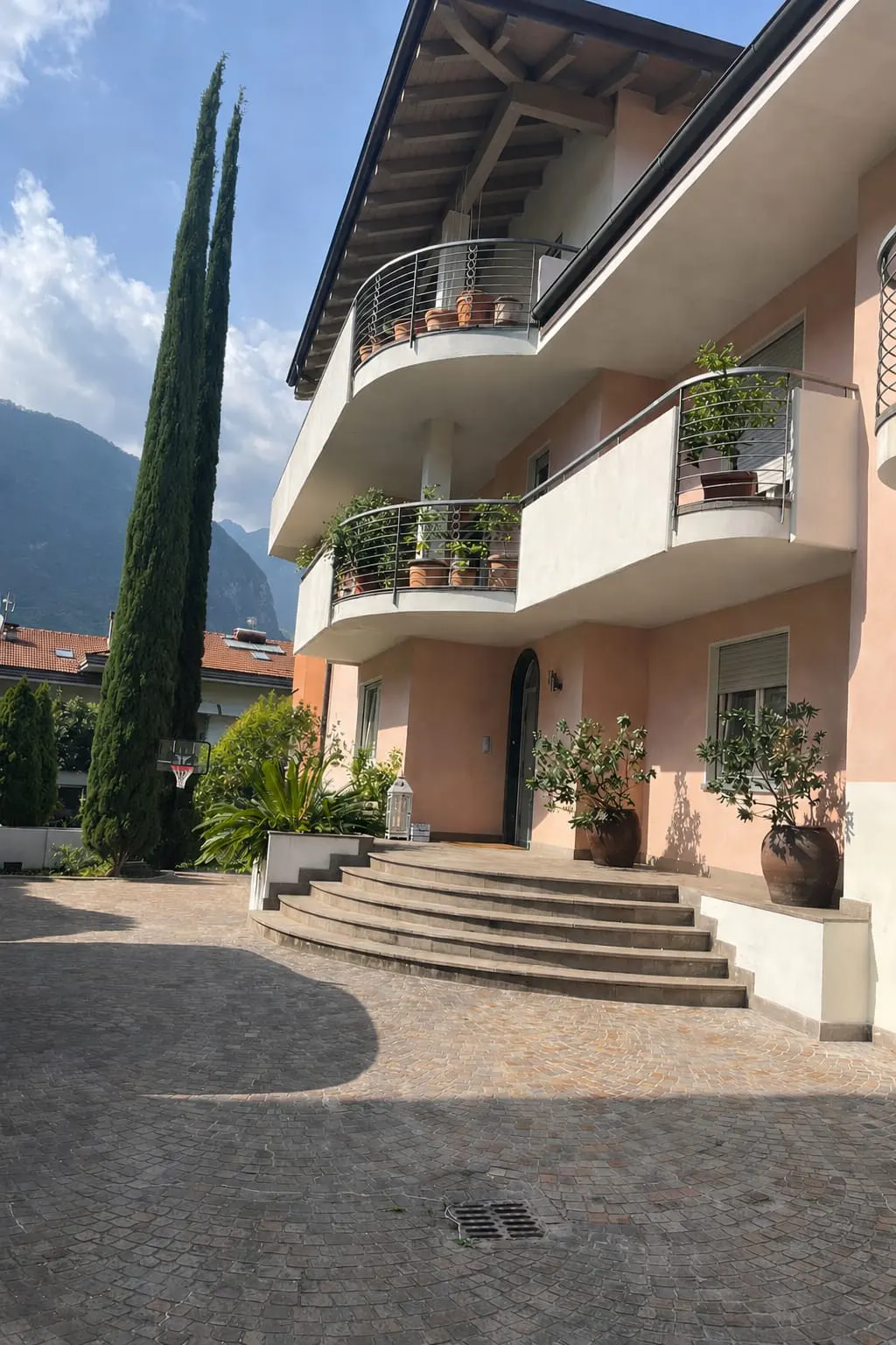 A peach-colored multi-story house with curved balconies, stone steps, and potted plants, set against a mountain backdrop.