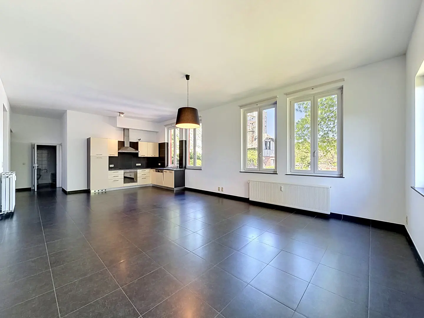 Bright, open-concept apartment with black tile floors, white walls, and a modern kitchen with light wood cabinets. Large windows offer natural light.