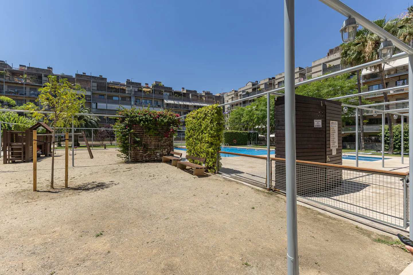 Outdoor community area with a sandy playground, benches, lush green privacy screens, and a swimming pool under blue sky.