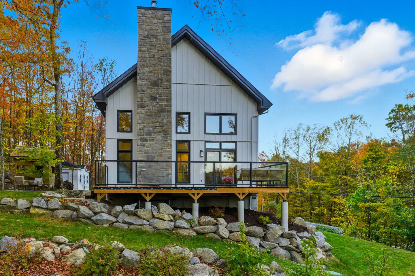 Modern white farmhouse with a stone chimney, black trim, and a wooden deck overlooking a green lawn and colorful fall foliage.