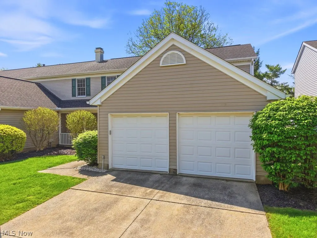 Attached two-car garage with white doors and tan siding. Green lawn and bushes in front of the house. Blue sky.