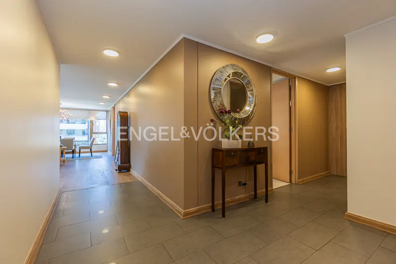 A hallway with gray tile floors, beige walls, and recessed lighting. A round mirror hangs above a wooden console table with flowers.