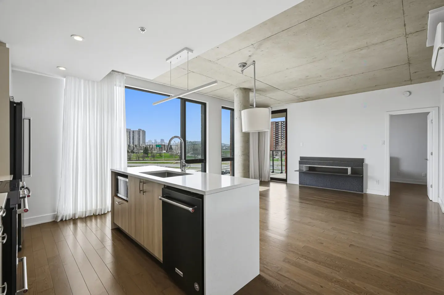 Modern condo interior with a kitchen island, hardwood floors, and a concrete ceiling. Large windows offer a city view.