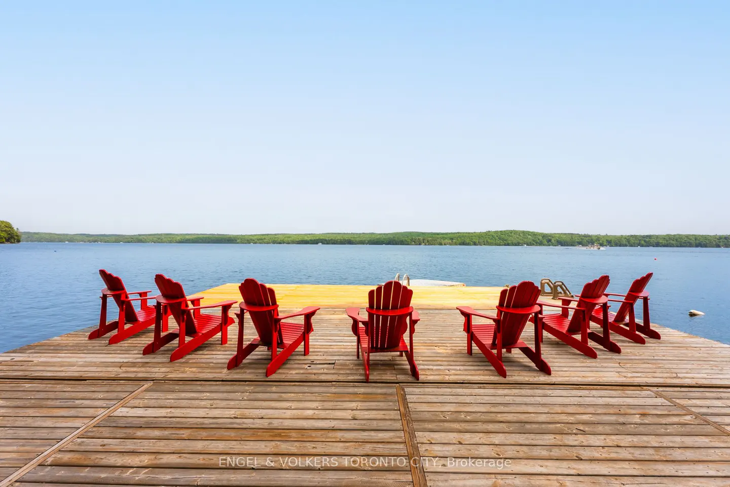 A wooden dock with eight red Adirondack chairs facing a blue lake and green trees under a clear sky.