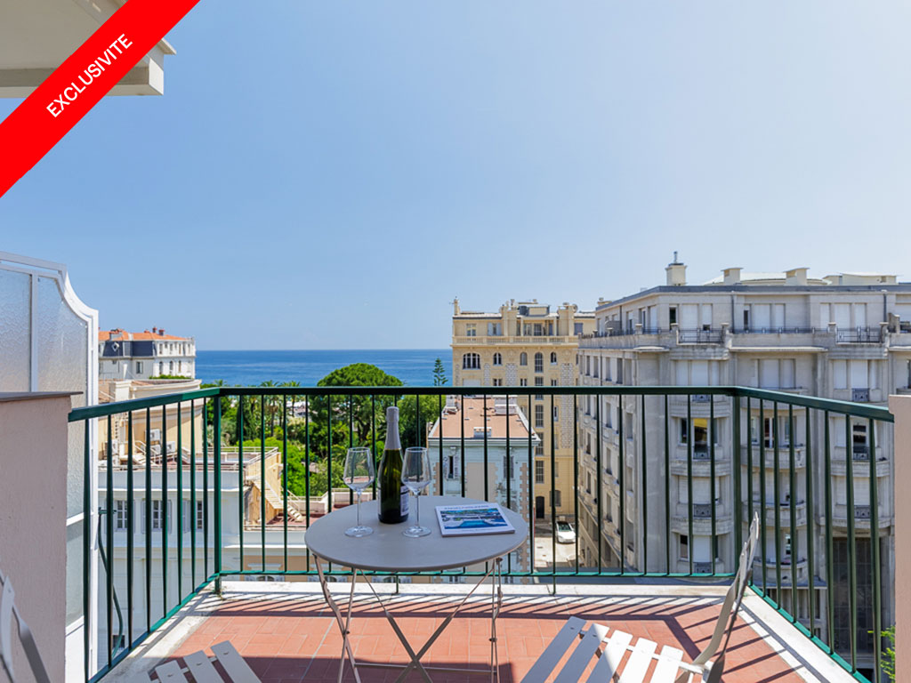 Balcony with a small table, two wine glasses, and a bottle, overlooking a city street and the blue sea under a clear sky.