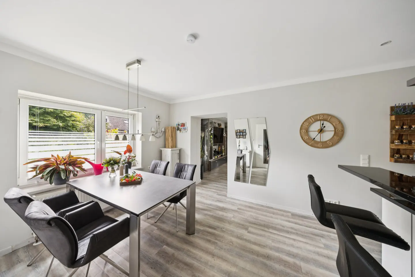 Bright dining area with a gray table, black chairs, and wood-look floors. A window with plants and a view outside adds natural light.