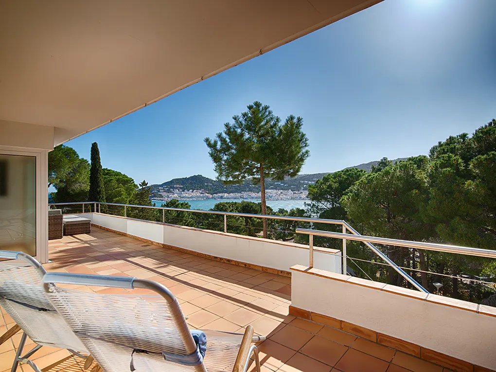 Balcony view with lounge chairs, terracotta tiles, and a railing overlooking trees, the ocean, and a distant town under a clear blue sky.