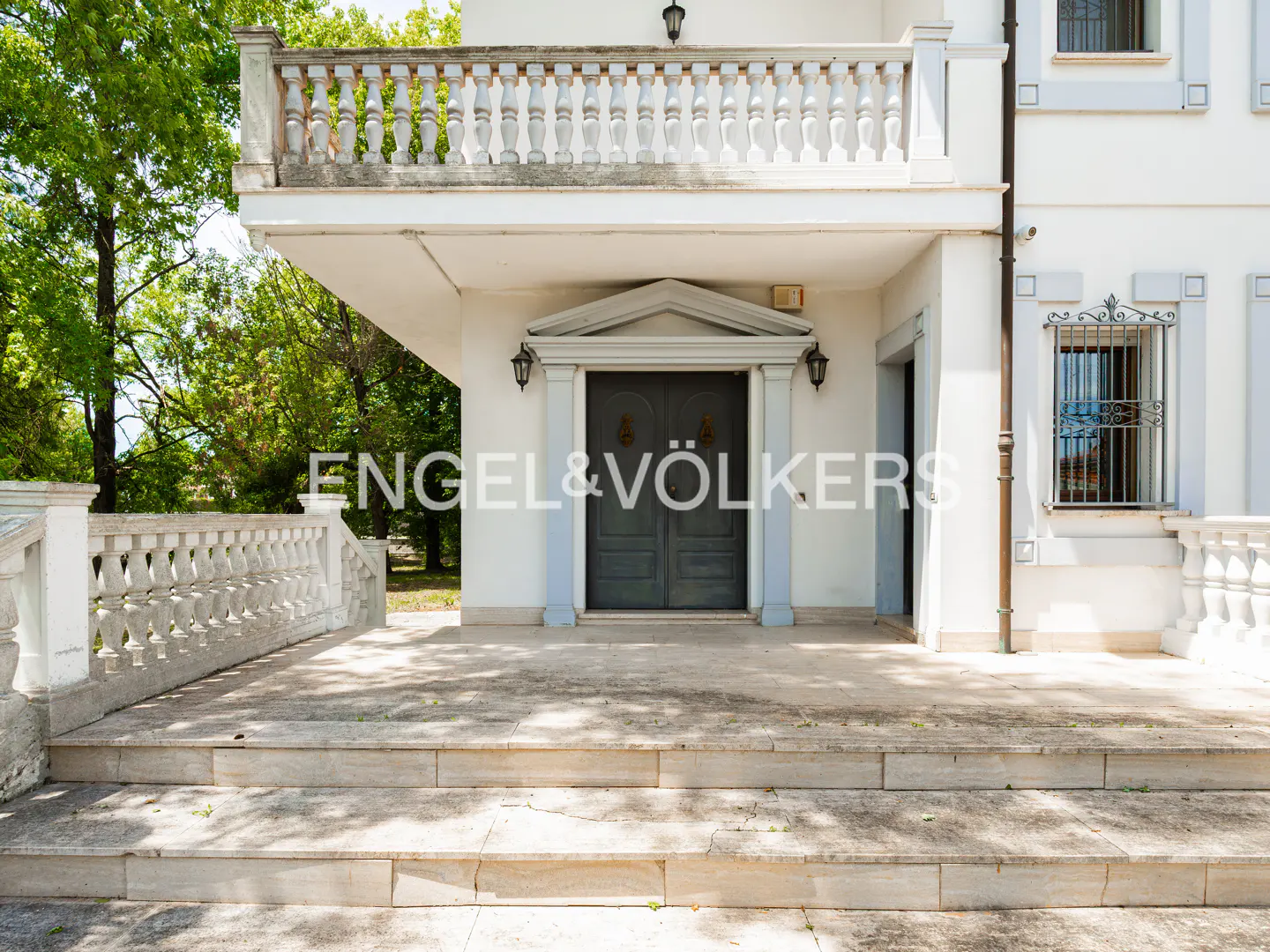 White stone steps lead to a grand entrance with dark double doors, a stone balcony above, and lush green trees nearby.