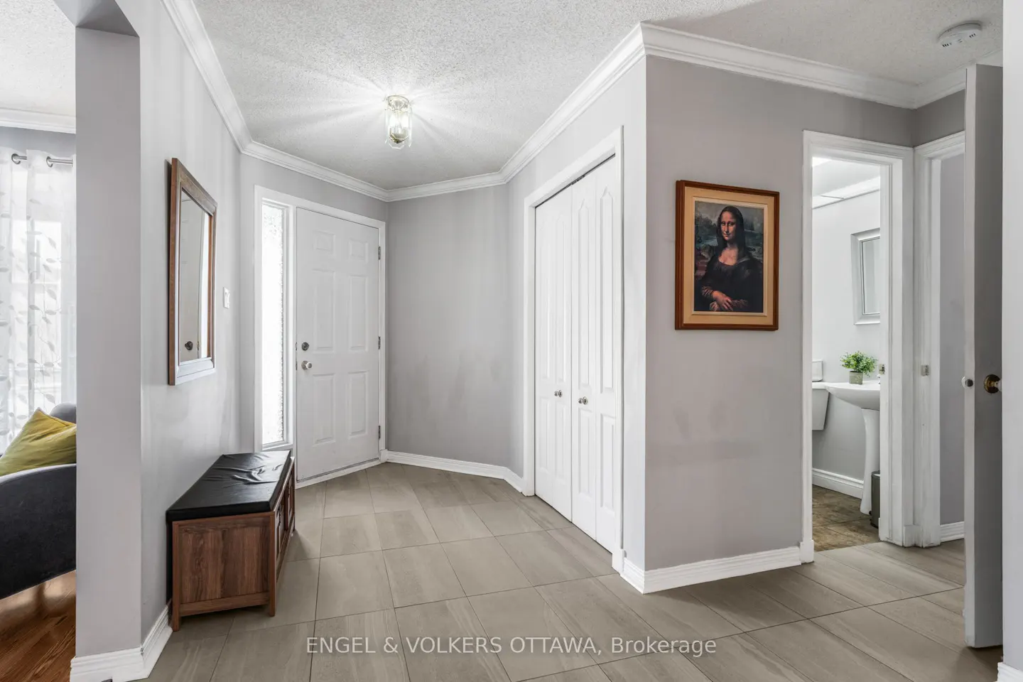 A bright foyer with gray walls, white trim, and tile floors. A white front door, closet, and bathroom are visible. A Mona Lisa print hangs on the wall.