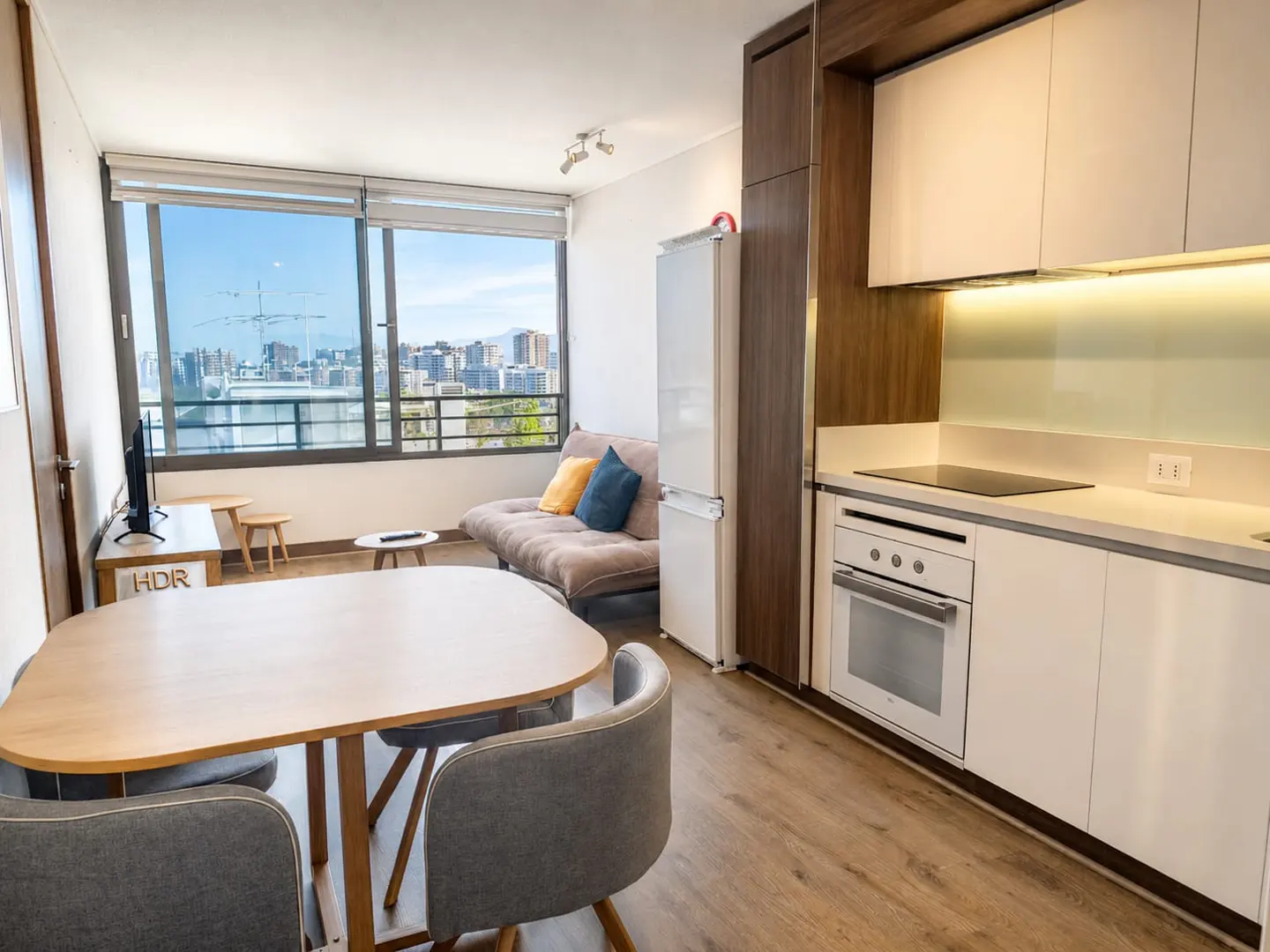 A modern apartment interior with a dining table, sofa, kitchen, and a large window with a city view.