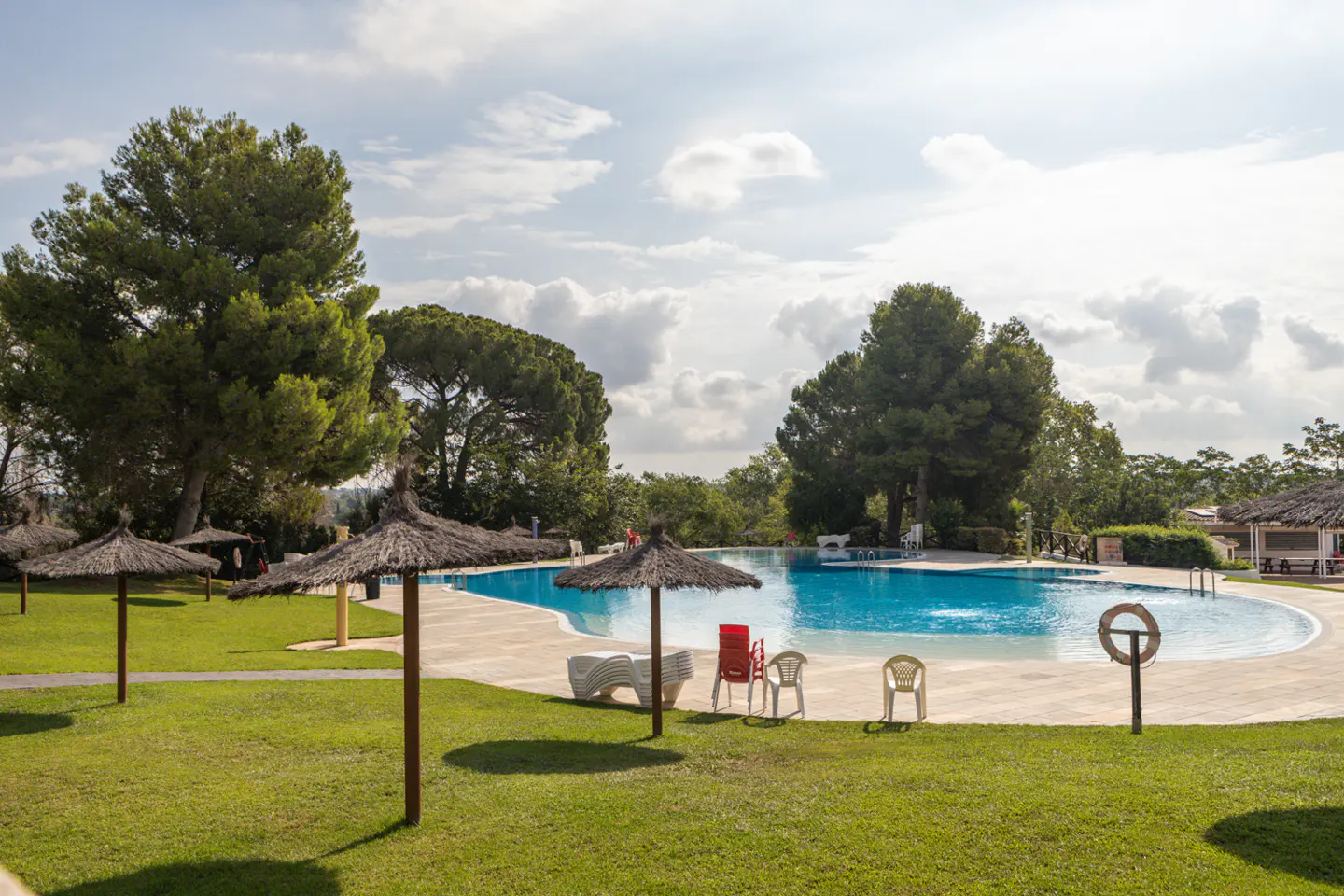 Outdoor pool with blue water, surrounded by green grass, trees, and straw umbrellas under a cloudy sky.