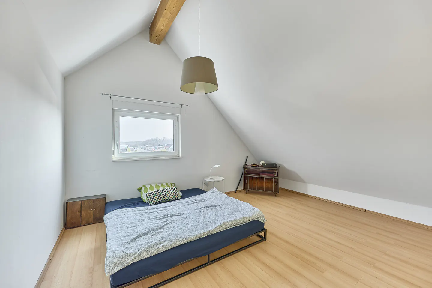 Attic bedroom with slanted white walls, wooden floors, a low bed with blue bedding, and a pendant light.